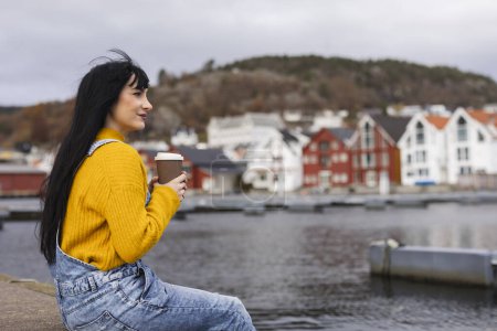 Eine junge Frau entspannt sich am Wasser und genießt ihren Kaffee mit einer malerischen norwegischen Stadt im Hintergrund.