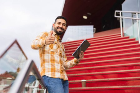An enthusiastic male entrepreneur gives a thumbs up, holding a tablet on red steps.