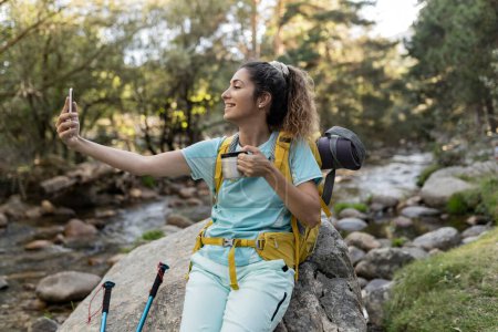 A vibrant young woman captures a selfie moment while enjoying a hot drink beside a babbling stream during a hiking break.