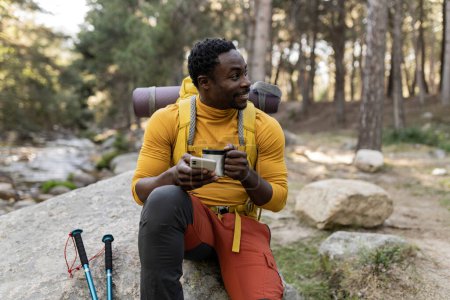 Ein zufriedener männlicher Wanderer sitzt auf einem Felsen, nippt am Kaffee und benutzt sein Smartphone in einer ruhigen Waldkulisse.