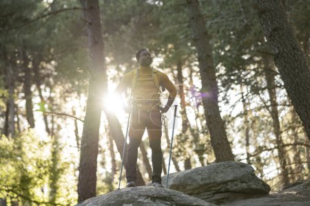 Téléchargez les photos : Un randonneur masculin se tient fièrement sur un rocher, se prélassant à la lumière du soleil qui traverse le couvert forestier dense. - en image libre de droit