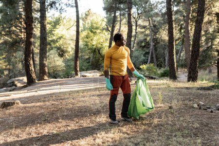 Un caminante masculino, equipado con guantes, lleva una bolsa de basura recogida, haciendo su parte para mantener la belleza natural del bosque.