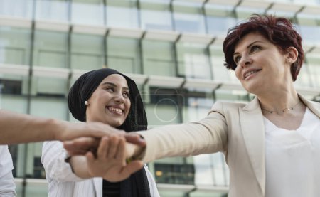 Two smiling female professionals from different cultural backgrounds joining hands in a gesture of unity and teamwork outside a modern office.