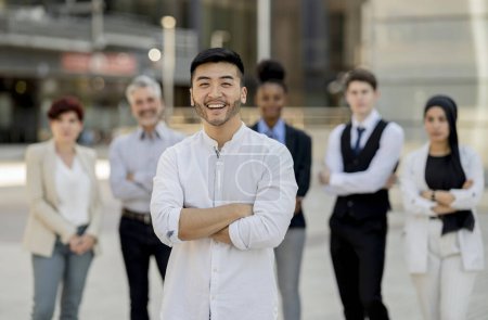 Hombre de negocios asiático sonriente con los brazos cruzados frente a un grupo diverso de profesionales, simbolizando el liderazgo en un entorno corporativo.