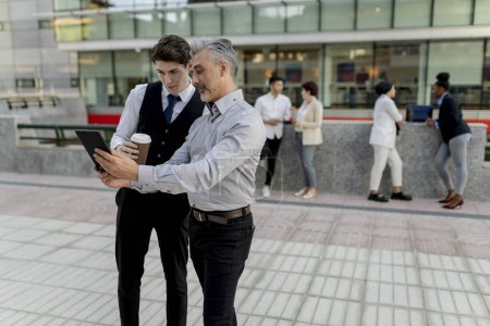 Homme d'affaires senior partageant des idées avec un jeune collègue masculin utilisant une tablette numérique, lors d'une réunion informelle à l'extérieur.