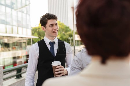 Un joven profesional masculino sonriendo mientras sostiene una taza de café y conversando con una colega, con edificios de oficinas en el fondo.