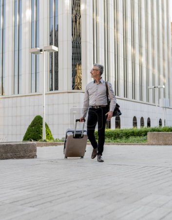 Mature professional man carrying a suitcase while walking briskly near modern office buildings, portraying a business travel scenario.