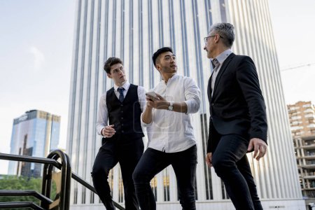 Drei Geschäftsleute unterschiedlichen Alters führen eine angeregte strategische Diskussion, bei der sie ihr Gespräch in einem städtischen Umfeld mit einem Smartphone erleichtern.