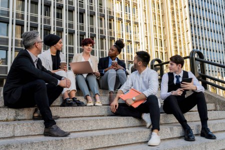 A dynamic group of multiethnic business professionals engaged in a collaborative meeting while seated on the steps outside a modern office building, using digital devices to enhance their discussion.