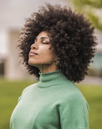 A confident young woman with a stunning afro hairstyle looks contemplative in a serene outdoor setting, wearing a vibrant green sweater.