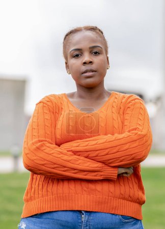A confident young woman poses outdoors, her expression serious and thoughtful, wearing a striking orange cable-knit sweater.