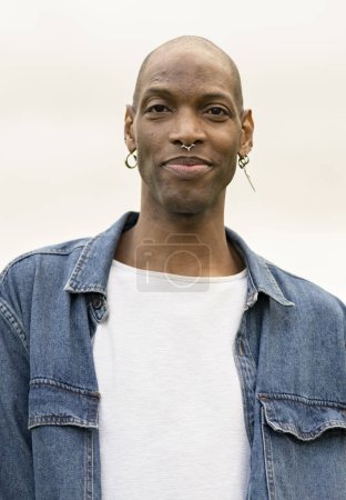 A portrait of a young man with a shaved head, showcasing unique jewelry including earrings and a nose ring, wearing a classic denim jacket.
