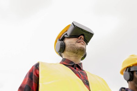 Low angle view of a young adult male construction worker wearing a VR headset, equipped with safety gear.