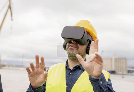 Männlicher Ingenieur mittleren Alters mit einem VR-Headset, gestikuliert beim Einsatz von Virtual-Reality-Technologie auf einer Baustelle.