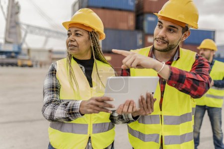 African American female engineer and Hispanic male coworker discuss over a tablet at a busy port.