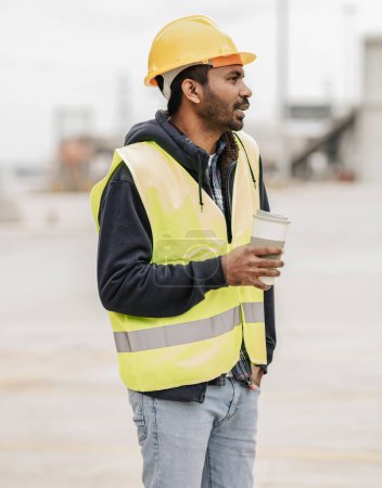 Young South Asian male construction worker holding a coffee cup, wearing a safety helmet and vest at a work site.