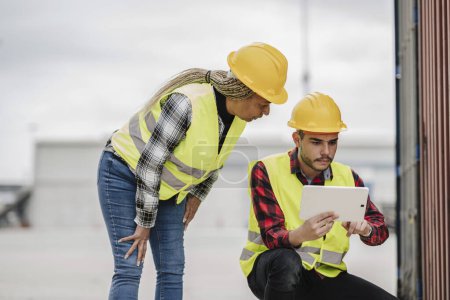 Focused African American woman and Hispanic man in safety gear inspecting project details on a digital tablet at an industrial location.