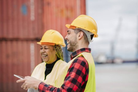African American woman and Hispanic man in hard hats and safety vests laughing together at an industrial site.