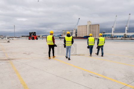 Groupe de travailleurs de la construction en engins de sécurité se dirigeant vers leur zone de travail dans un port occupé, mettant en valeur le travail d'équipe et l'environnement industriel.