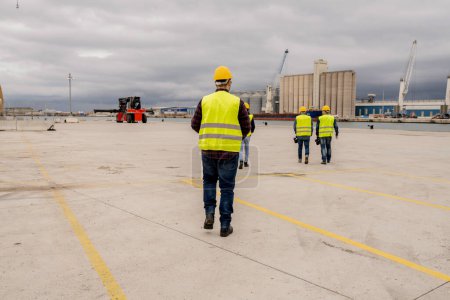 Un ouvrier de la construction dirige un groupe de collègues à travers un port spacieux, mettant l'accent sur le leadership et le travail d'équipe dans un cadre industriel.