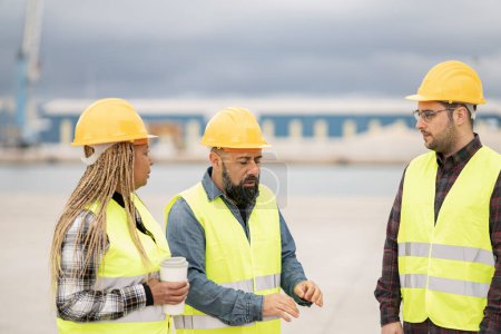 Diverse construction team engaged in a discussion at a port, with an African American woman, Middle Eastern man, and Caucasian man in safety gear.