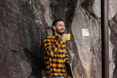 Ein fröhlicher junger hispanischer Mann steht mit Laptop und Kaffee entspannt an einer Felswand und genießt eine Pause im Freien..