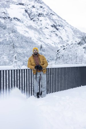 Photographer wearing a yellow beanie and warm jacket, holding a camera, stands in a snowy landscape with a mountain background.