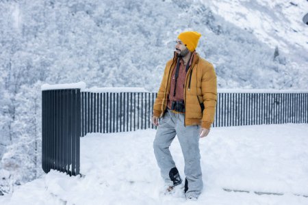 Ein bärtiger Mann in gelber Mütze und isolierter Jacke geht durch tiefen Schnee, Kamera in der Hand, in einer atemberaubenden, verschneiten Bergkulisse.