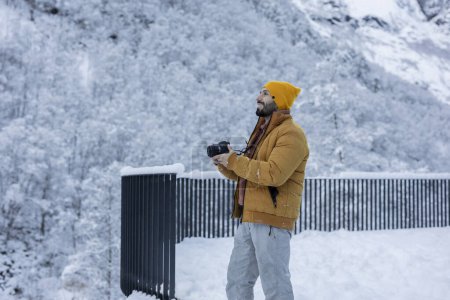 Joyful photographer in a yellow beanie and warm jacket captures the beauty of a snowy mountain landscape with his camera.