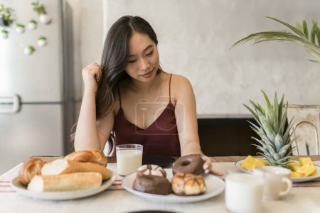 An Asian woman in her twenties sits at a breakfast table, surrounded by an assortment of pastries, fresh fruit, and milk.