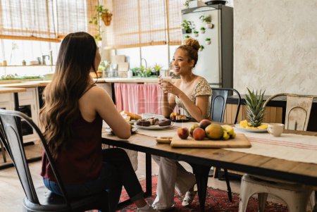 Two young women share a joyful conversation over a breakfast spread that includes coffee, fresh fruits, and pastries in a sunny, rustic kitchen.