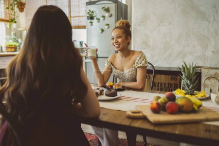 A mixed-race woman shares a smile and a conversation with her Asian friend over a breakfast of pastries and fresh fruit in a bohemian-style kitchen.