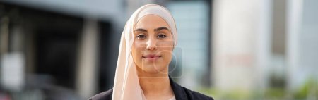 Heitere und doch selbstbewusste muslimische Geschäftsfrau im Hijab, die in einer modernen städtischen Landschaft steht und ihre berufliche Stärke und Eleganz widerspiegelt.