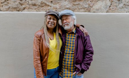Charming and diverse senior couple smiling warmly while sharing a close embrace, highlighting their affection and stylish urban wear against a rustic wall backdrop.