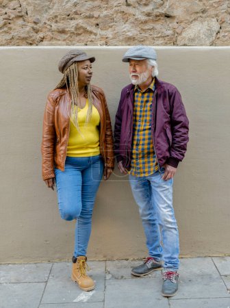 Un couple de personnes âgées diversifié partageant un moment de conversation contre un mur urbain rustique, mettant en valeur leur style à la mode et détendu.