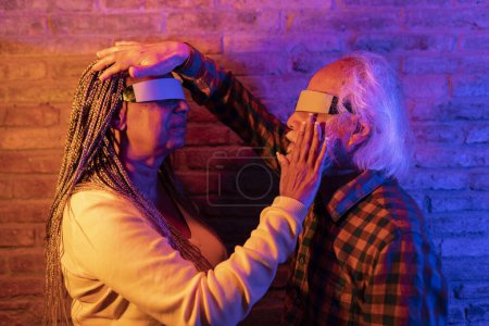 Elderly couple exploring virtual reality, hands touching, surrounded by colorful lighting.