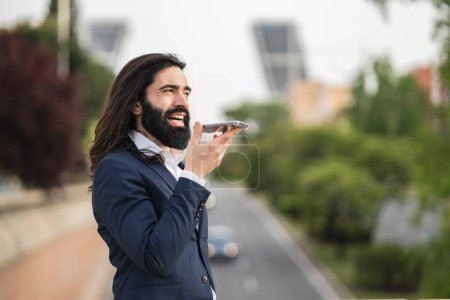 Energetic young businessman speaking into his phone using voice commands, actively engaging with technology in an urban setting.
