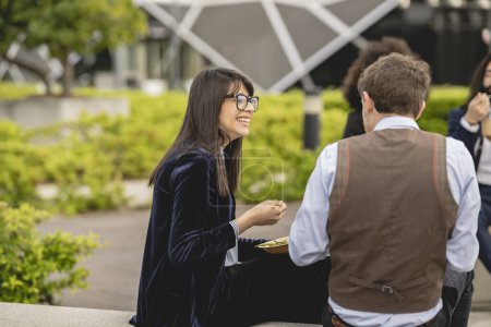 Jeunes professionnels souriants profitant d'une conversation animée autour d'un déjeuner dans un parc urbain.