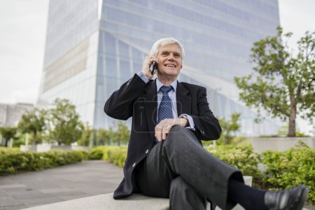 Sonriente hombre de negocios de edad avanzada en un traje haciendo una llamada telefónica mientras está sentado al aire libre.