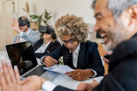 Una empresaria animada con el pelo rizado coordina una discusión enfocada en la tecnología mientras sus colegas se involucran con la realidad virtual en la oficina.
