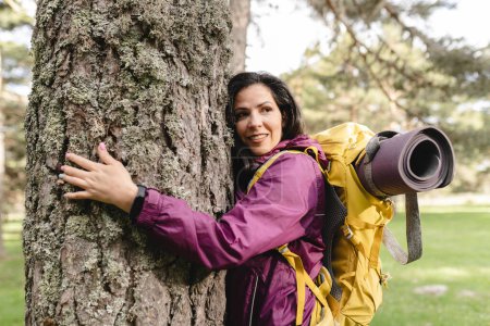 A female nature photographer with a backpack hugs a large tree in the forest, showcasing her connection to nature.
