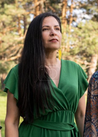A thoughtful woman in a vibrant green dress with yellow earrings gazes into the distance amidst a serene forest backdrop.