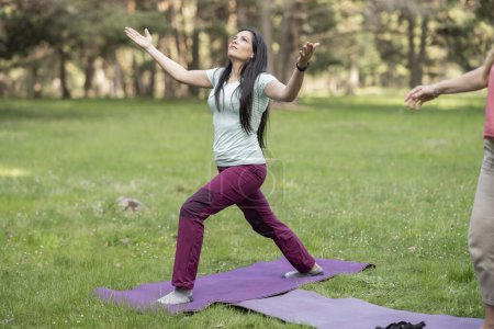 Eine Frau genießt die Ruhe eines Waldes, während sie Yoga praktiziert und auf einer violetten Matte auf einer Rasenfläche posiert.