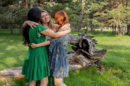 Three women share a heartfelt hug in a sunlit forest, expressing joy and friendship surrounded by nature.