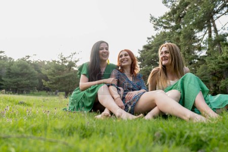Three women enjoy a laughter-filled moment as they sit closely together in a lush forest meadow, showcasing their close bond.