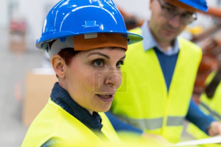 Close-up of a middle-aged female engineer in a hard hat and high-visibility vest, engaged in a project discussion.