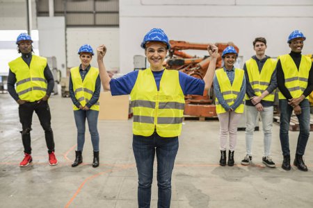 Ingeniera femenina de mediana edad en chaleco de alta visibilidad y sombrero duro que lidera con confianza a su diverso equipo en una instalación industrial.