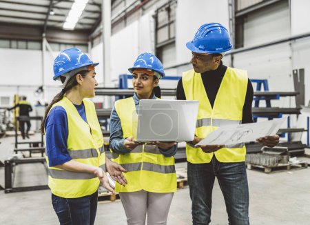 Group of engineers wearing safety gear discussing plans in an industrial warehouse, using a laptop.
