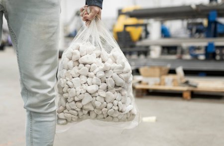 Close-up of a worker holding a mesh bag filled with white industrial materials in a factory.