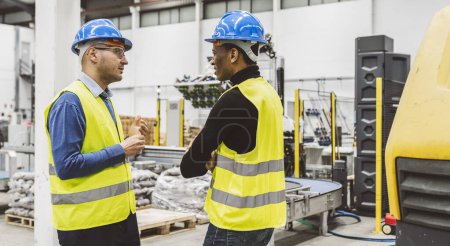 Two engineers wearing hard hats and high-visibility vests engaged in a discussion inside an industrial facility.